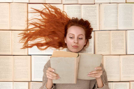 A young girl with a book in her hands lies on the background of books. Redhead girl dressed in beige sweater lies on books. Distance learning.の写真素材