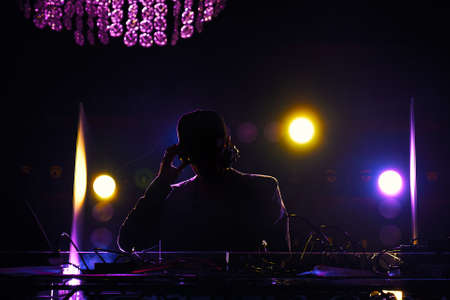 A disc jockey at the turntable. DJ mixing tracks on a mixer in a nightclub.DJ silhouette over foggy...