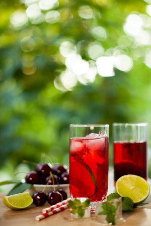 Two glasses of cold fruit drink, served with cherry, mint, lime, ice, and a straw. Summer relaxation in the backyard shade. Selective focus, copy space.の写真素材
