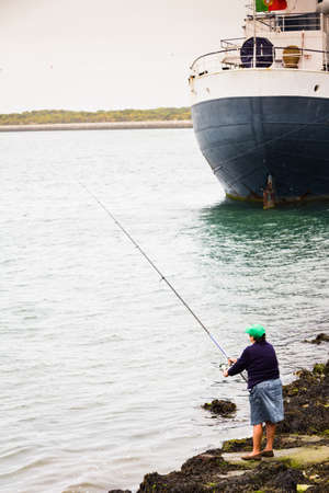 Local mature woman with green cap fishing near the big cargo shipの写真素材