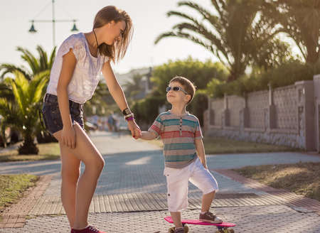 Beautiful young girl and cute kid walking with a skateboards outdoor on summerの写真素材