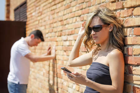 Angry woman in focus looking phone and frustrated man posing his hands on the wall backgroundの写真素材