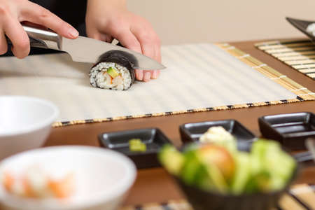 Closeup of woman chef cutting japanese sushi rolls with rice, avocado and shrimps on nori seaweed sheetの写真素材