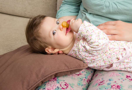 Adorable baby playing with the pacifier lying in her mother legs at home