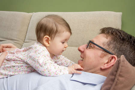 Portrait of cute baby playing with her happy father lying down over his belly in a sofa at home