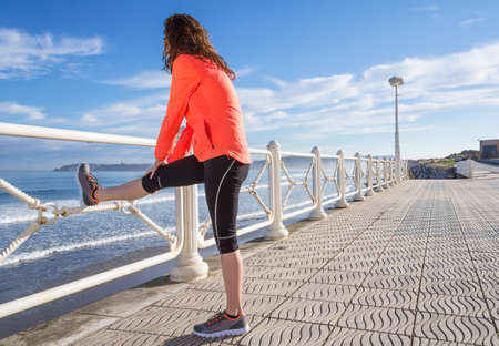 Athletic young girl warming before running in the beach promenade on a summer morning over blue cloudy sky backgroundの写真素材