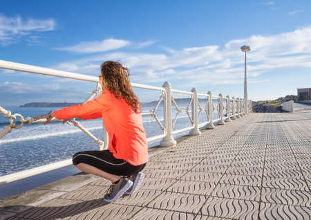 Athletic young girl warming before running in the beach promenade on a summer morning over blue cloudy sky backgroundの写真素材