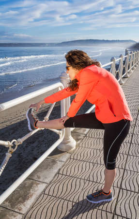 Athletic young girl warming before running in the beach promenade on a summer morning over blue...