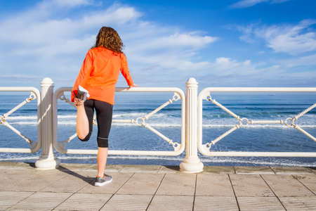 Athletic young girl warming before running in the beach promenade on a summer morning over blue cloudy sky backgroundの写真素材