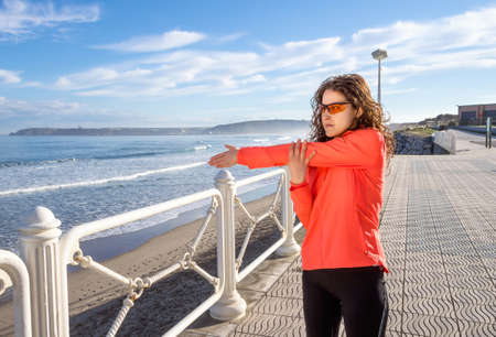 Athletic young girl warming before running in the beach promenade on a summer morning over blue...