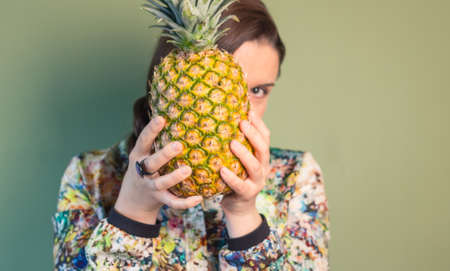 Colorful fashion girl portrait holding a pineapple in front of her face over green backgroundの写真素材