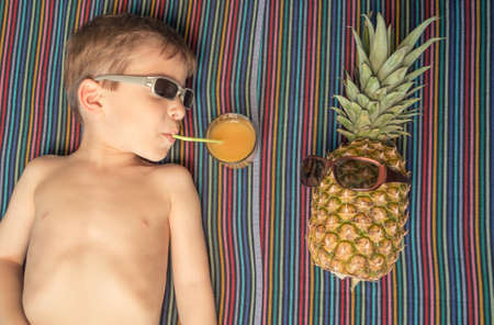 Cute happy child drinking juice and pineapple with sunglasses sunbathing lying down over a striped towel on summerの写真素材