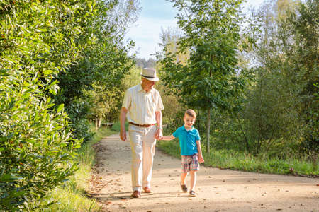 Front view of grandfather with hat and grandchild walking on a nature pathの写真素材