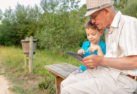 Grandchild teaching to his grandfather to use a electronic tablet on a park bench. Focus on grandfather. Generation values concept.の写真素材