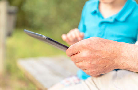 Closeup of grandfather hands using a electronic tablet with his granchild on a park bench....