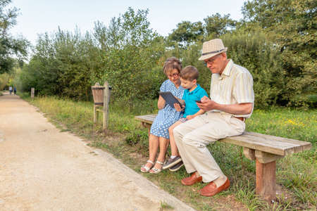Grandchild teaching to his grandmother to use a electronic tablet on a park bench. Generation values concept.の写真素材
