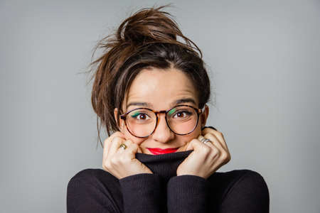 Portrait of real young girl with glasses and red lips taking her black neck sweater with the hands over a gray backgroundの写真素材