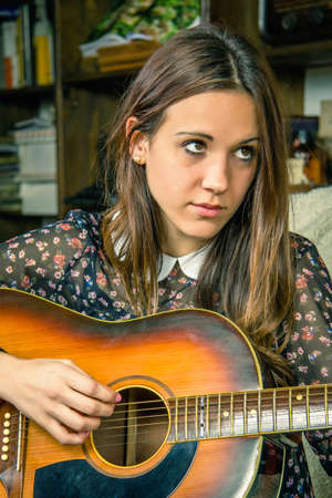 Portrait of beautiful young hipster girl playing acoustic guitar sitting in a sofa at home. Retro vintage colors editionの写真素材
