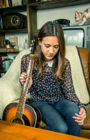 Beautiful young hipster girl with acoustic guitar sitting in a sofa at home. Retro vintage colors editionの写真素材