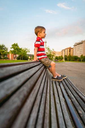 Portrait of boy with short pants sitting on the top of wooden bench park relaxing in a bored summer dayの写真素材