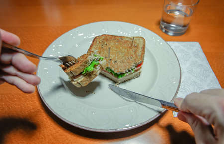 Closeup of man hands cutting sandwich with a fork and knife served in a plate over a wooden tableの写真素材