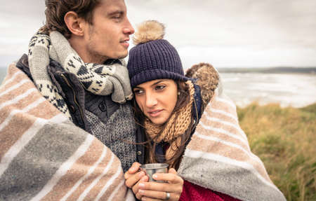 Closeup of young beautiful couple embracing under blanket in a cold day with sea and dark cloudy sky on the backgroundの写真素材