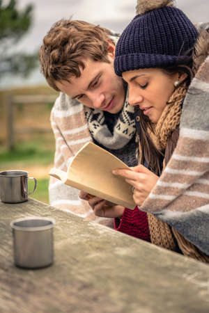 Closeup of young beautiful couple under blanket reading book in a cold day with sea and dark cloudy sky on the backgroundの写真素材