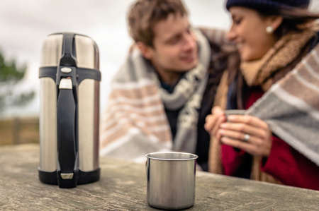 Closeup of metalic cup and thermos with hot beverage in a wooden table with young couple under blanket blurred on the backgroundの写真素材