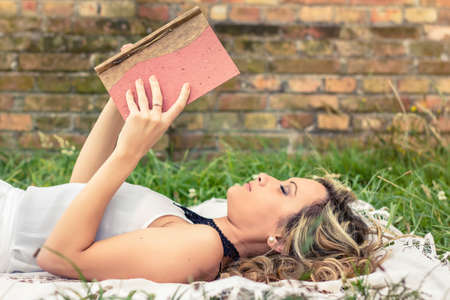 Portrait of romantic young woman reading a book lying over the grass. Relax outdoor time concept.