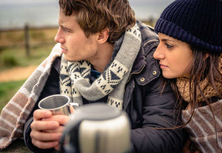 Closeup of young beautiful couple under blanket having hot beverage in a cold day with dark cloudy sky on the backgroundの写真素材