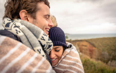 Closeup of young happy couple laughing under blanket in a cold day with sea and dark cloudy sky on the backgroundの写真素材