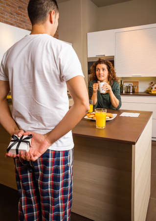 Back view of man hiding a gift box behind his back and his surprised girlfriend waiting while having breakfast on the home kitchenの写真素材