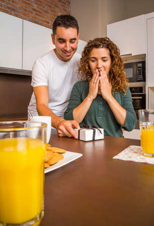 Young man giving a gift box to his surprised girlfriend while having breakfast in the home kitchenの写真素材