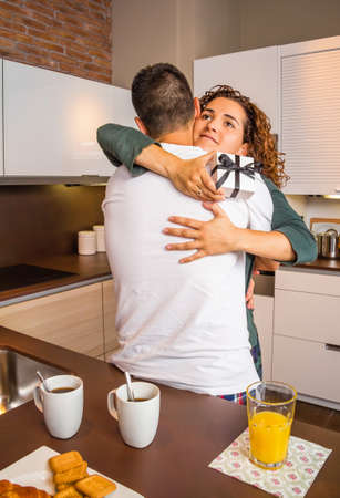 Young girl with gift box in the hands embracing to her boyfriend for a beautiful surprise while having breakfast on the home kitchenの写真素材