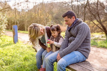 Man and woman playing with a little girl sitting on the center of a wooden bench in the park. Family...