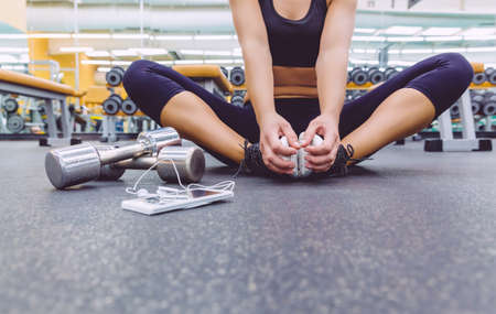 Closeup of sporty woman sitting on the floor of fitness center with dumbbells and smartphone with...