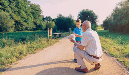 Grandfather showing his hat to grandchild over a nature path background. Two different generations concept.の写真素材