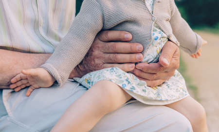 Closeup of baby girl sitting over legs of senior man outdoors. Two different generations concept.の写真素材