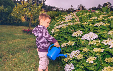 Portrait of happy cute boy watering flowers in the garden with a blue watering can