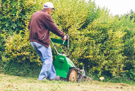 Back view of senior man with cap mowing lawn with a lawnmower machine in a field