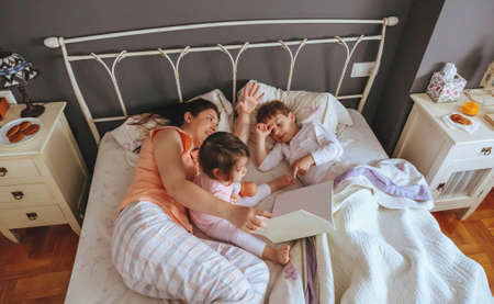 Top view of mother reading story book to her daughter and son kids lying in the bed. Weekend family...