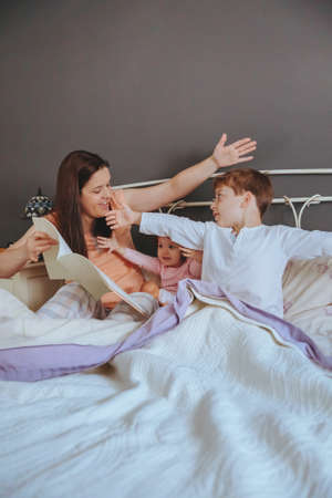 Portrait of mother reading story book to her daughter and son kids sitting in the bed. Weekend...
