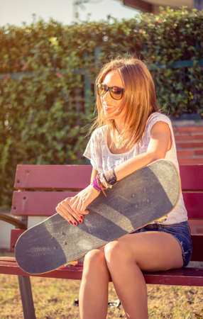 Portrait of beautiful young girl with short shorts and skateboard sitting on bench in a summer day. Warm tones edition.の写真素材