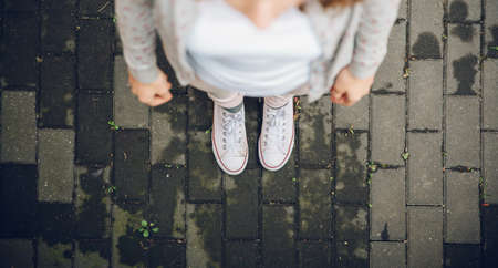 Top view of young trendy woman feet in white sneakers over a gray street paving stone backgroundの写真素材