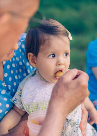 Closeup of senior man feeding with fruit puree to adorable baby girl sitting over a senior woman in a bench outdoors. Grandparents and grandchildren lifestyle concept.の写真素材