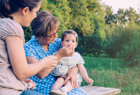 Closeup of senior woman feeding with fruit puree to adorable baby girl sitting in a bench outdoors....