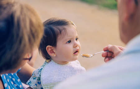 Back view of senior man feeding with fruit puree to adorable baby girl sitting over a senior woman outdoors. Grandparents and grandchildren lifestyle concept.の写真素材