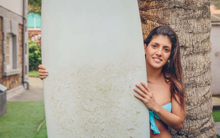 Closeup of beautiful surfer woman with bikini holding a surfboard in front of palm treeの写真素材