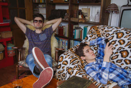 Young man looking smartphone lying on sofa and friend relaxing in rocking chair in a bored day at home. Teenager leisure time concept.の写真素材