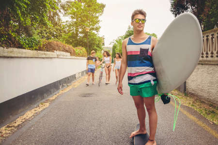 Young man riding on a skate and holding surfboard with group of friends having fun in the background...